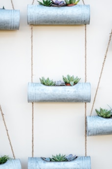 Several metal planters are vertically arranged and suspended by ropes against a white wall. Each planter contains a variety of small succulents with shades of green and hints of purple.
