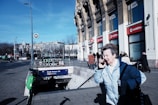 A city street scene featuring a person in a light blue coat using a phone while walking past a metro station entrance. The building in the background has several storefronts, including Vodafone, and the sky is clear and blue. The street is moderately busy with pedestrians and some parked scooters near the entrance labeled 'Deák Ferenc tér'.
