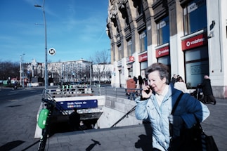 A city street scene featuring a person in a light blue coat using a phone while walking past a metro station entrance. The building in the background has several storefronts, including Vodafone, and the sky is clear and blue. The street is moderately busy with pedestrians and some parked scooters near the entrance labeled 'Deák Ferenc tér'.