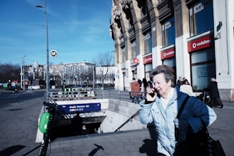 A city street scene featuring a person in a light blue coat using a phone while walking past a metro station entrance. The building in the background has several storefronts, including Vodafone, and the sky is clear and blue. The street is moderately busy with pedestrians and some parked scooters near the entrance labeled 'Deák Ferenc tér'.