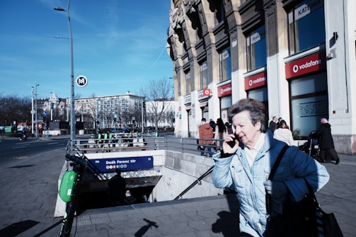 A city street scene featuring a person in a light blue coat using a phone while walking past a metro station entrance. The building in the background has several storefronts, including Vodafone, and the sky is clear and blue. The street is moderately busy with pedestrians and some parked scooters near the entrance labeled 'Deák Ferenc tér'.