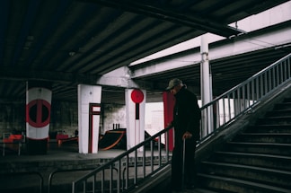 An elderly person using a cane is descending stairs in a dimly lit urban environment under a bridge. The area features concrete pillars painted with abstract designs in red and black. Some skateboard ramps and urban furniture are visible in the background.