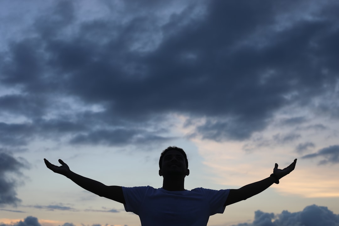 man in white t-shirt raising his hands under cloudy sky during daytime