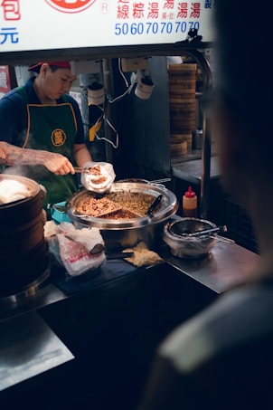 A street food vendor is preparing food at a stall. The vendor is wearing a cap and green apron, using a scoop to fill a wrap with a meat mixture from a large metal container. Various tools and ingredients are around the workstation, including steamer baskets and a squeeze bottle.