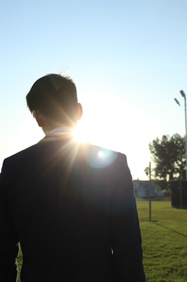 A person in a dark suit is standing outdoors, with the sun shining directly behind them, creating a silhouette effect. The background is a clear sky with some trees on the right side.