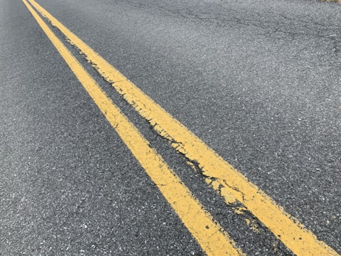 A close-up view of an asphalt road with two parallel yellow lines running down the center. The lines are slightly worn and cracked, with visible texture on the road surface.