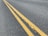 A close-up view of an asphalt road with two parallel yellow lines running down the center. The lines are slightly worn and cracked, with visible texture on the road surface.