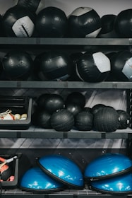 Several shelves filled with various gym equipment, including black medicine balls, blue half-balance balls, and other exercise tools. The equipment is neatly organized, with larger items placed on the lower shelves and smaller weights at the top.