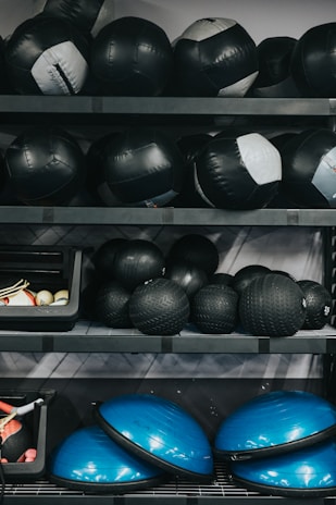 Several shelves filled with various gym equipment, including black medicine balls, blue half-balance balls, and other exercise tools. The equipment is neatly organized, with larger items placed on the lower shelves and smaller weights at the top.