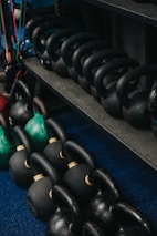A collection of black and green kettlebells is neatly arranged on a blue carpeted floor and a dark metal shelf. The kettlebells vary in size and are equipped with sturdy handles, some displaying signs of wear.