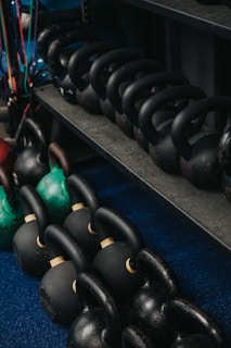 A collection of black and green kettlebells is neatly arranged on a blue carpeted floor and a dark metal shelf. The kettlebells vary in size and are equipped with sturdy handles, some displaying signs of wear.