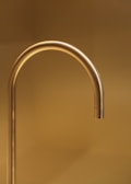 A sleek, gold-colored faucet extends with a graceful curve against a warm, golden-hued background. The simplicity and elegance of the design are accentuated by the soft lighting, creating a luxurious and minimalistic aesthetic.