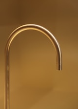A sleek, gold-colored faucet extends with a graceful curve against a warm, golden-hued background. The simplicity and elegance of the design are accentuated by the soft lighting, creating a luxurious and minimalistic aesthetic.
