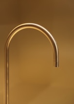 A sleek, gold-colored faucet extends with a graceful curve against a warm, golden-hued background. The simplicity and elegance of the design are accentuated by the soft lighting, creating a luxurious and minimalistic aesthetic.