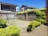 A small, neatly maintained garden with green shrubs and a grass lawn is enclosed by a stone wall. A metal swing is situated in the middle of the area. Two residential buildings with stone facades and tiled roofs are visible in the background under a clear blue sky.