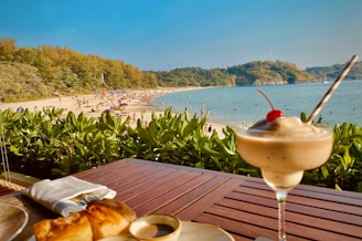 A beach scene with people enjoying the sun and sea, surrounded by lush greenery. In the foreground, there is a table with a dessert topped with a cherry and a croissant placed on a wooden surface.