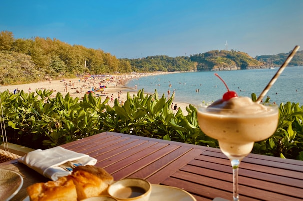 A beach scene with people enjoying the sun and sea, surrounded by lush greenery. In the foreground, there is a table with a dessert topped with a cherry and a croissant placed on a wooden surface.