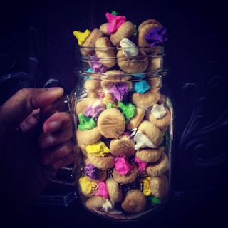 A glass jar filled with round biscuits topped with colorful icing in shades of pink, green, purple, white, blue, and yellow, held by a hand against a dark background.