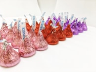 A group of chocolate candies wrapped in colorful foil are arranged in rows. The wrappers are in shades of pink, red, and purple, with each candy having a small paper tag protruding from the top.