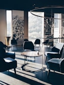 A stylish, modern lounge area with velvet armchairs and round wooden tables. Large windows offer a view of the outdoors, complemented by vintage black and white photographs on the walls. Soft, natural light casts long shadows across the floor, enhancing the elegant and serene ambiance.