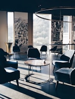 A stylish, modern lounge area with velvet armchairs and round wooden tables. Large windows offer a view of the outdoors, complemented by vintage black and white photographs on the walls. Soft, natural light casts long shadows across the floor, enhancing the elegant and serene ambiance.