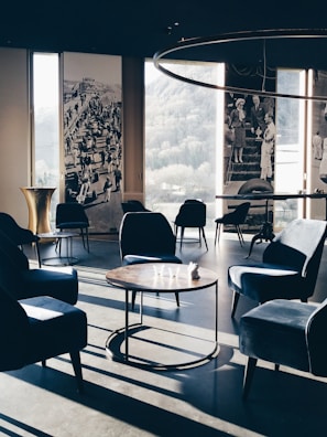 A stylish, modern lounge area with velvet armchairs and round wooden tables. Large windows offer a view of the outdoors, complemented by vintage black and white photographs on the walls. Soft, natural light casts long shadows across the floor, enhancing the elegant and serene ambiance.