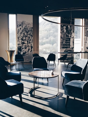 A stylish, modern lounge area with velvet armchairs and round wooden tables. Large windows offer a view of the outdoors, complemented by vintage black and white photographs on the walls. Soft, natural light casts long shadows across the floor, enhancing the elegant and serene ambiance.