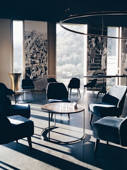 A stylish, modern lounge area with velvet armchairs and round wooden tables. Large windows offer a view of the outdoors, complemented by vintage black and white photographs on the walls. Soft, natural light casts long shadows across the floor, enhancing the elegant and serene ambiance.