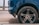 The image depicts the rear side of a blue car with a focus on the tire and rim. The tire shows some wear and is situated on a city street with a concrete surface visible. Part of the car body and wheel arch can also be seen.