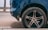 The image depicts the rear side of a blue car with a focus on the tire and rim. The tire shows some wear and is situated on a city street with a concrete surface visible. Part of the car body and wheel arch can also be seen.