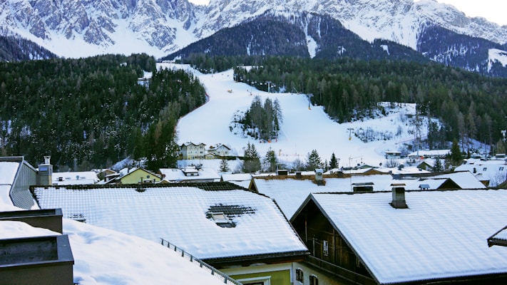 A snowy landscape featuring a small village with snow-covered rooftops is set against a backdrop of dense pine forests and towering, snow-capped mountains. A ski slope runs through the forest, marked by ski tracks and ski lifts. The scene captures a serene winter day with fresh snowfall, creating a picturesque alpine setting.