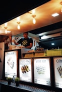 A food vendor is preparing meals inside a well-lit food stall. Brightly illuminated signs display various dish names and prices, including Tauhu Bakar, Pepes Ayam, and Satar Ikan. Decorative lights hang from the ceiling above the vendor.