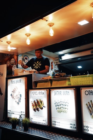 A food vendor is preparing meals inside a well-lit food stall. Brightly illuminated signs display various dish names and prices, including Tauhu Bakar, Pepes Ayam, and Satar Ikan. Decorative lights hang from the ceiling above the vendor.