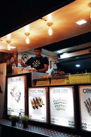 A food vendor is preparing meals inside a well-lit food stall. Brightly illuminated signs display various dish names and prices, including Tauhu Bakar, Pepes Ayam, and Satar Ikan. Decorative lights hang from the ceiling above the vendor.