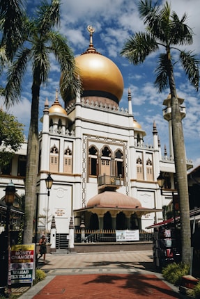 A grand mosque with a large golden dome and ornate architectural details. The structure is surrounded by palm trees, with a clear blue sky above. The signs on the street indicate services like money exchange and textiles.