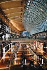 A spacious and modern shopping mall interior with a high, arched glass ceiling allowing natural light to illuminate the area. The space is divided into multiple levels with various retail stores visible along the walkways. The floors are shiny and well-polished, reflecting light, with several people walking around, adding a sense of bustling activity.