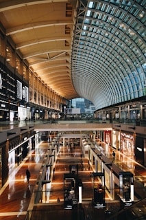 A spacious and modern shopping mall interior with a high, arched glass ceiling allowing natural light to illuminate the area. The space is divided into multiple levels with various retail stores visible along the walkways. The floors are shiny and well-polished, reflecting light, with several people walking around, adding a sense of bustling activity.