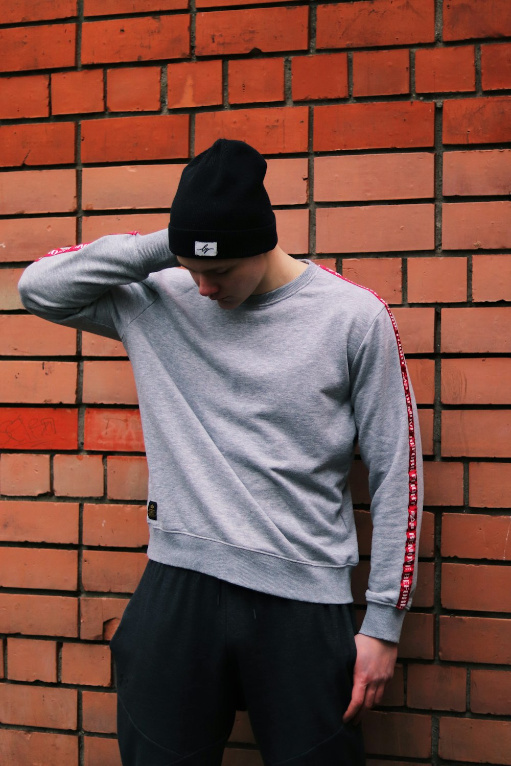 man in gray sweater and black cap standing beside red brick wall