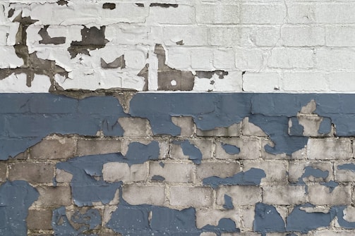 A brick wall with peeling paint, revealing layers of white, gray, and dark blue colors. The surface shows a mix of rough textures and exposed bricks, with old and worn paint giving it a weathered look.