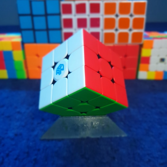 A Rubik's cube is centrally positioned, partially solved with visible red, white, and green faces. The cube is balanced on a stand, and in the background, there are several other cubes in various sizes and color patterns arranged on a blue surface.