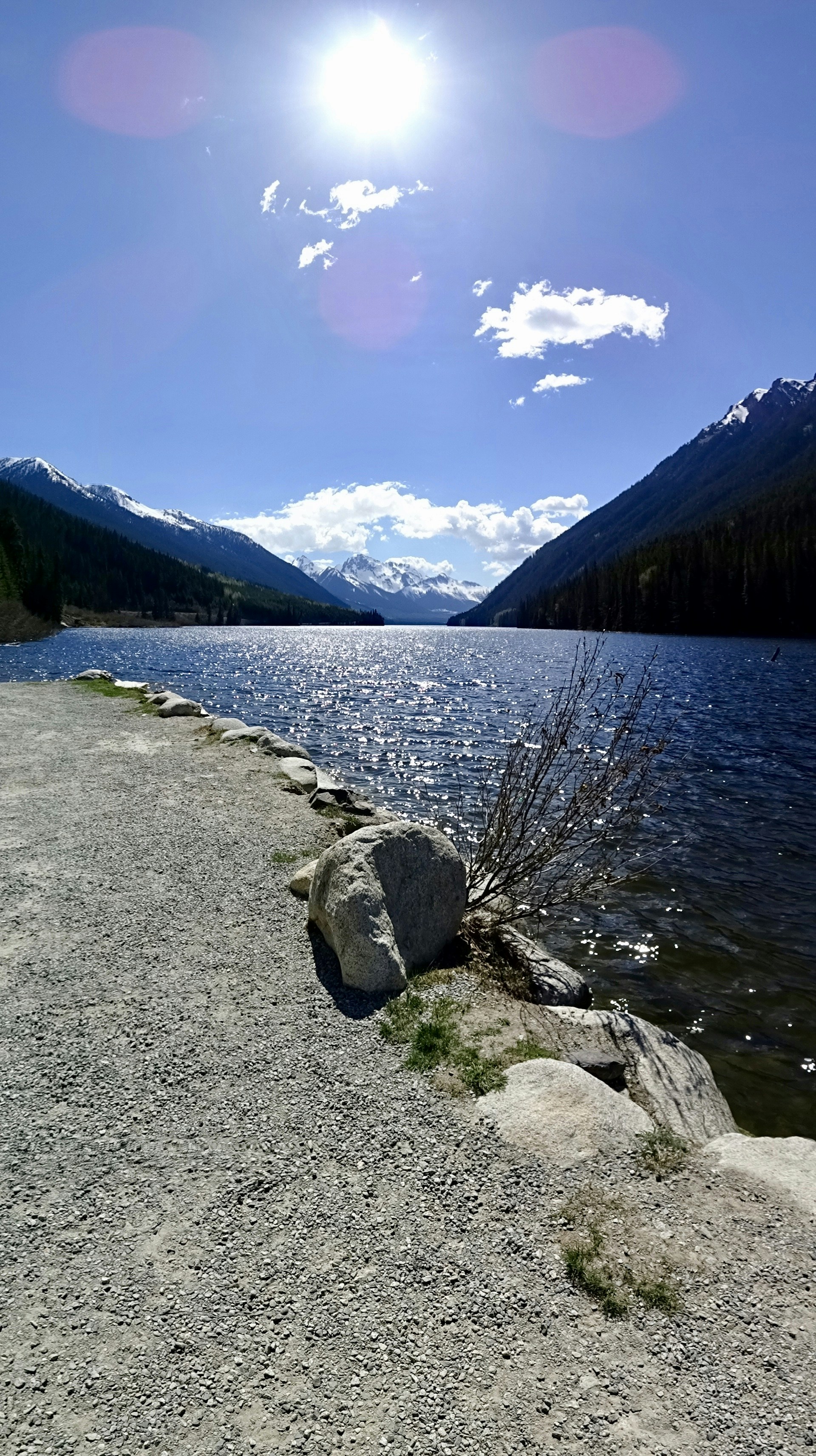 Sunlit alpine lake framed by snow-capped peaks, with a gravel path and rocks along the shore.
