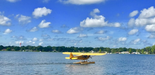 A yellow seaplane is floating on a calm body of water. In the background, there are trees and houses lining the shore under a bright blue sky with scattered white clouds.