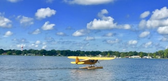 A yellow seaplane is floating on a calm body of water. In the background, there are trees and houses lining the shore under a bright blue sky with scattered white clouds.