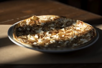 A pizza on a white plate with a topping of caramelized onions and diced cheese, set on a wooden table. The light is casting shadows, creating a warm and cozy atmosphere.