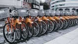 A row of rental bicycles with orange frames and silver baskets parked in an urban setting. The bikes are neatly aligned on a paved surface, with a backdrop of buildings and commercial signs.