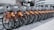 A row of rental bicycles with orange frames and silver baskets parked in an urban setting. The bikes are neatly aligned on a paved surface, with a backdrop of buildings and commercial signs.