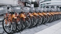 A row of rental bicycles with orange frames and silver baskets parked in an urban setting. The bikes are neatly aligned on a paved surface, with a backdrop of buildings and commercial signs.