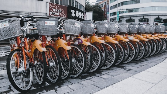 A row of rental bicycles with orange frames and silver baskets parked in an urban setting. The bikes are neatly aligned on a paved surface, with a backdrop of buildings and commercial signs.