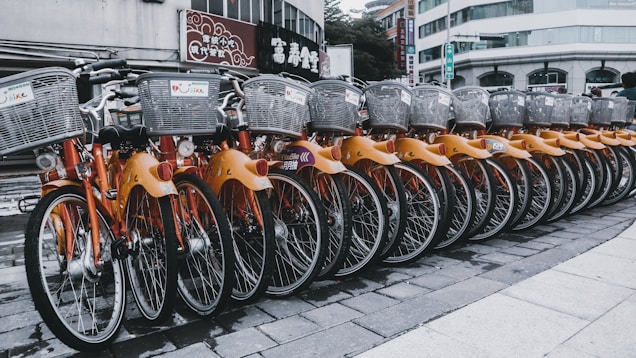 A row of rental bicycles with orange frames and silver baskets parked in an urban setting. The bikes are neatly aligned on a paved surface, with a backdrop of buildings and commercial signs.