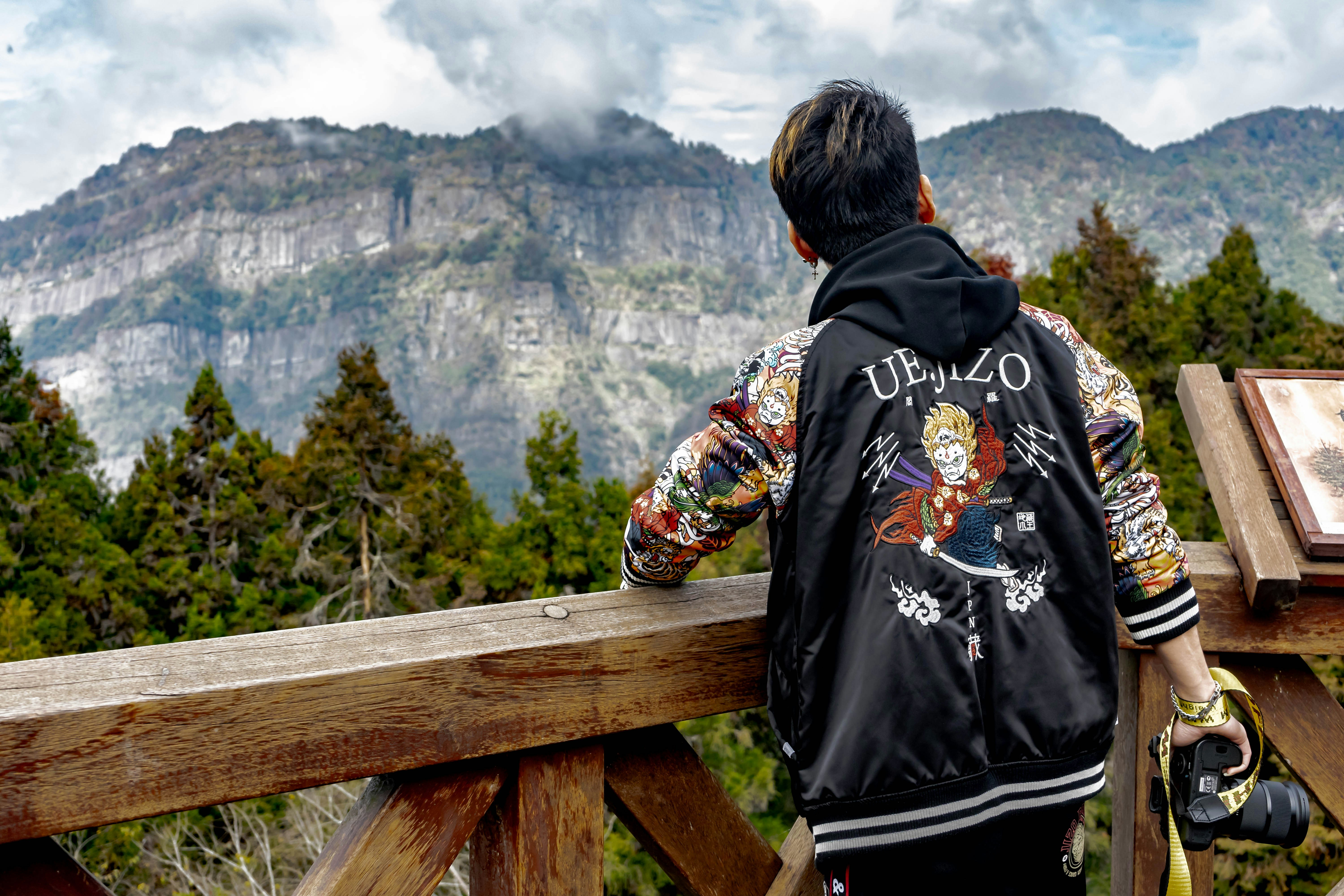 man in black and red hoodie sitting on brown wooden fence looking at green mountains during